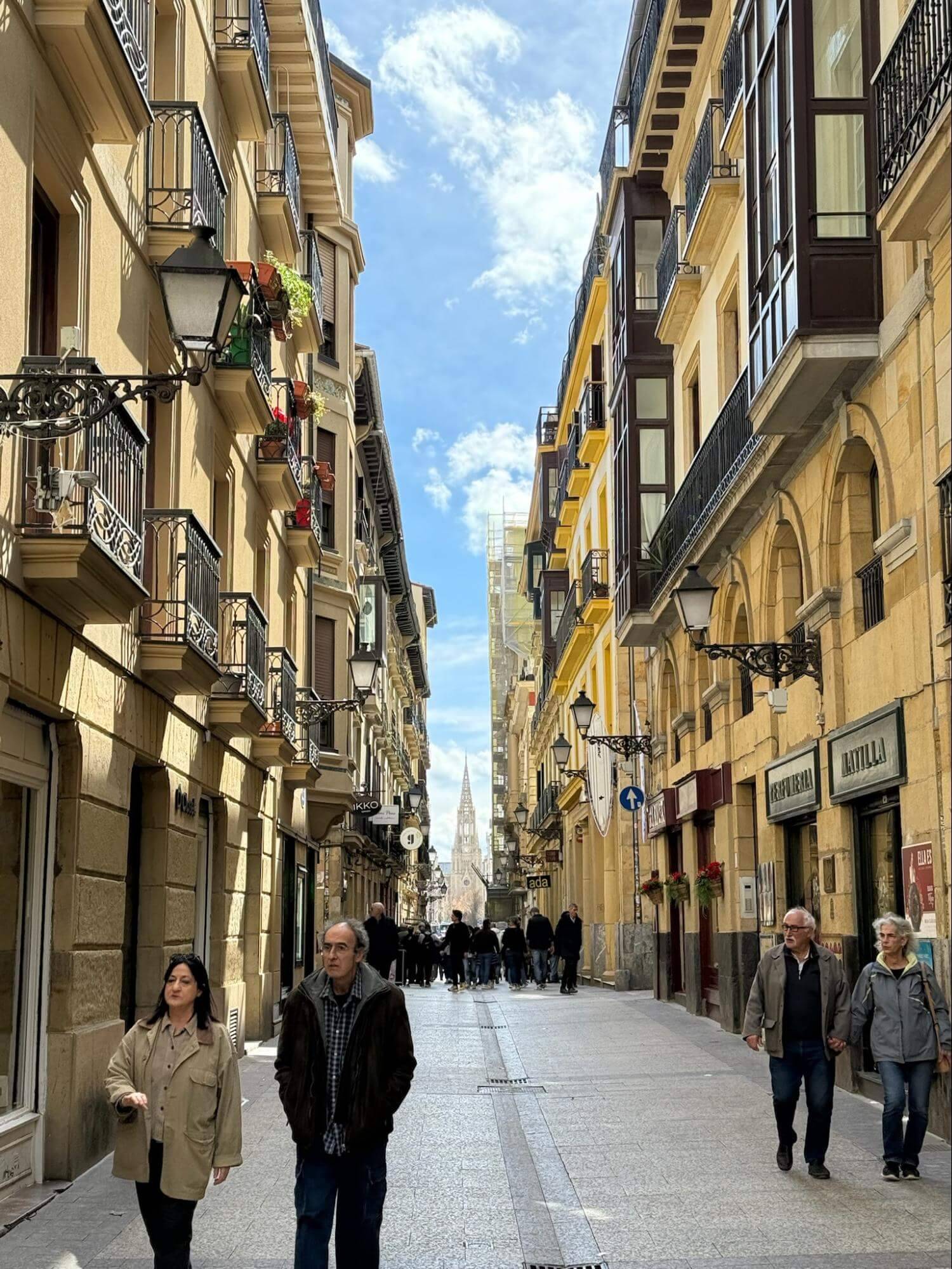 A bustling city street lined with charming buildings, balconies adorned with flowers, and a glimpse of a steeple in the distance under a bright sky.