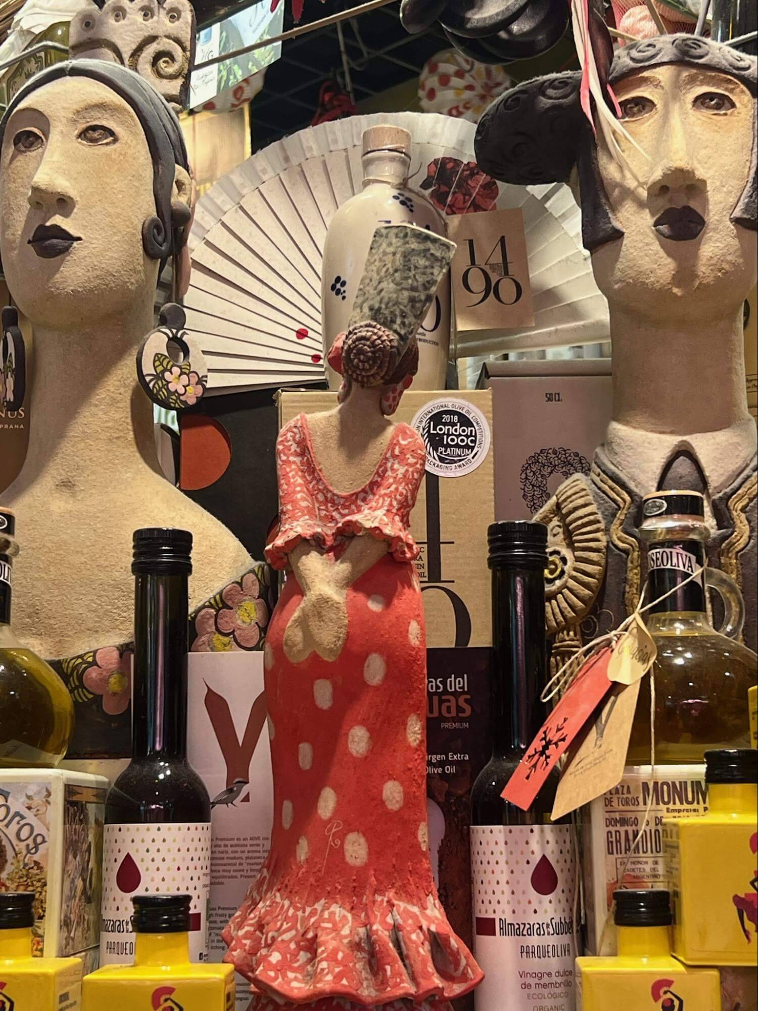 A colorful clay figurine in a polka-dot dress stands amidst various bottles and a decorative fan, showcasing a vibrant collection.