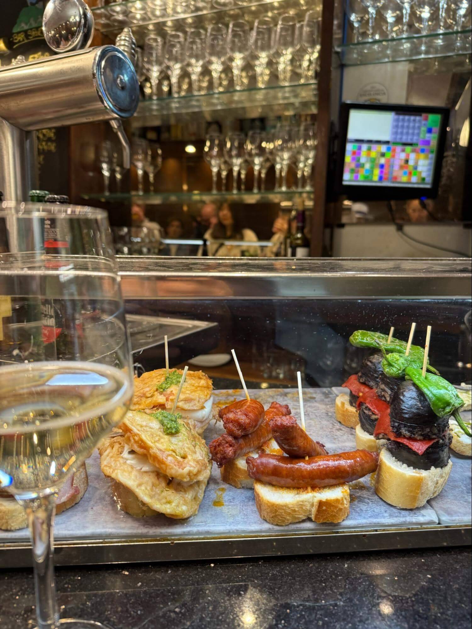A close-up of a variety of delicious tapas on small breads, with a glass of white wine in the foreground and bar ambiance in the background.