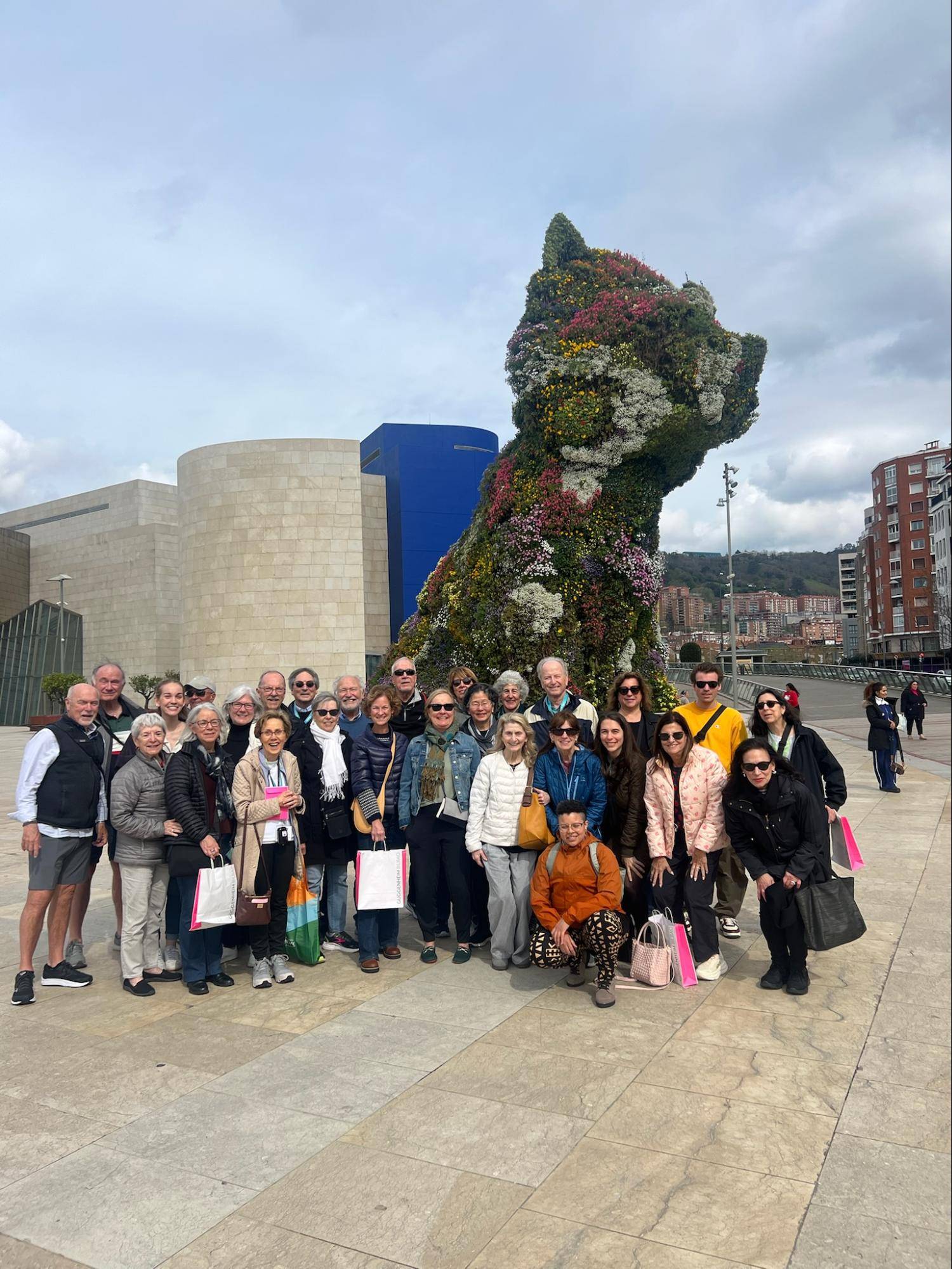 A group of tourists posing in front of a large floral sculpture of a dog in a city square, with modern buildings in the background.