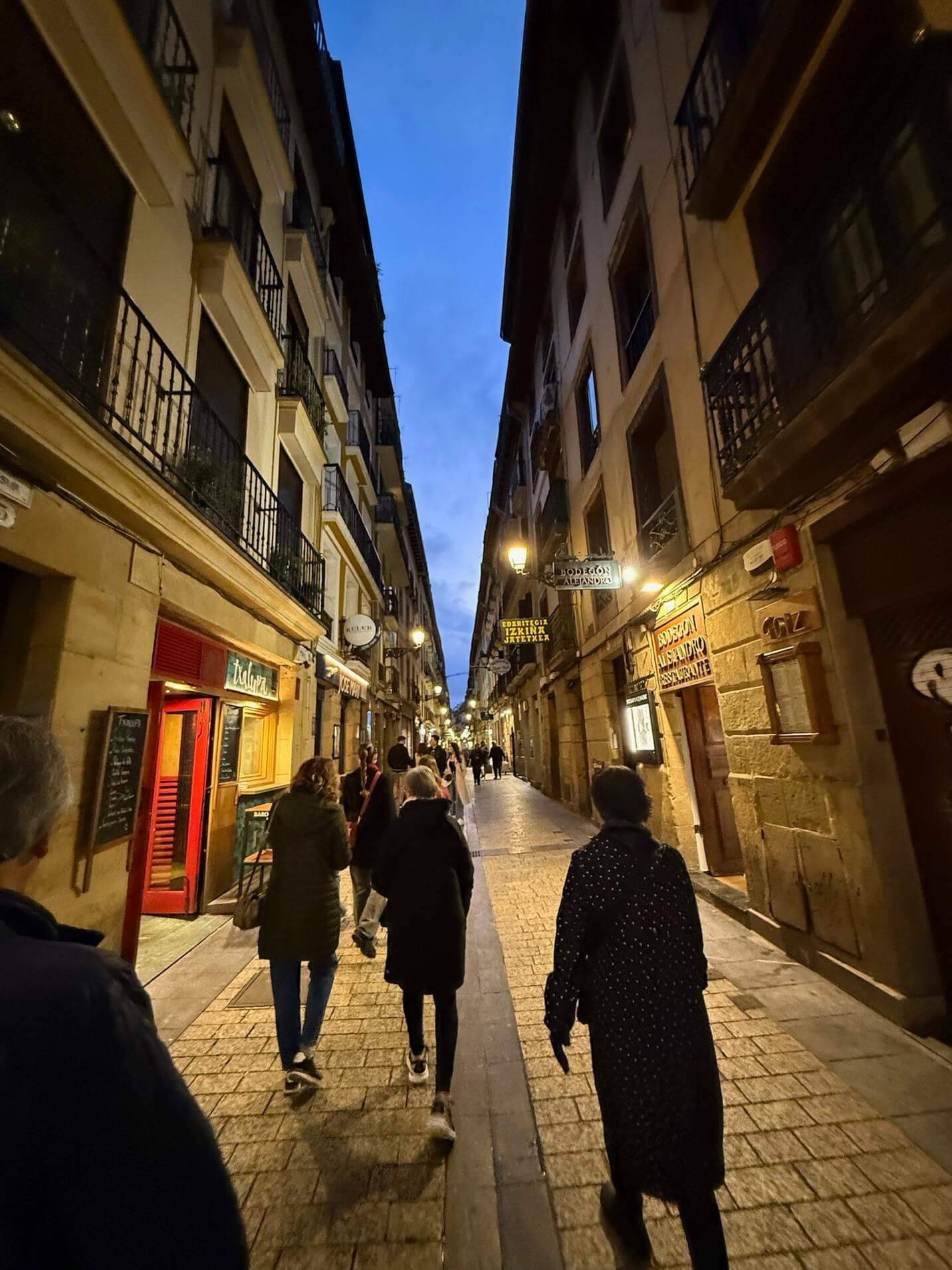 A narrow, cobblestone street lined with buildings, as people stroll under a dusky blue sky, illuminated by street lamps.