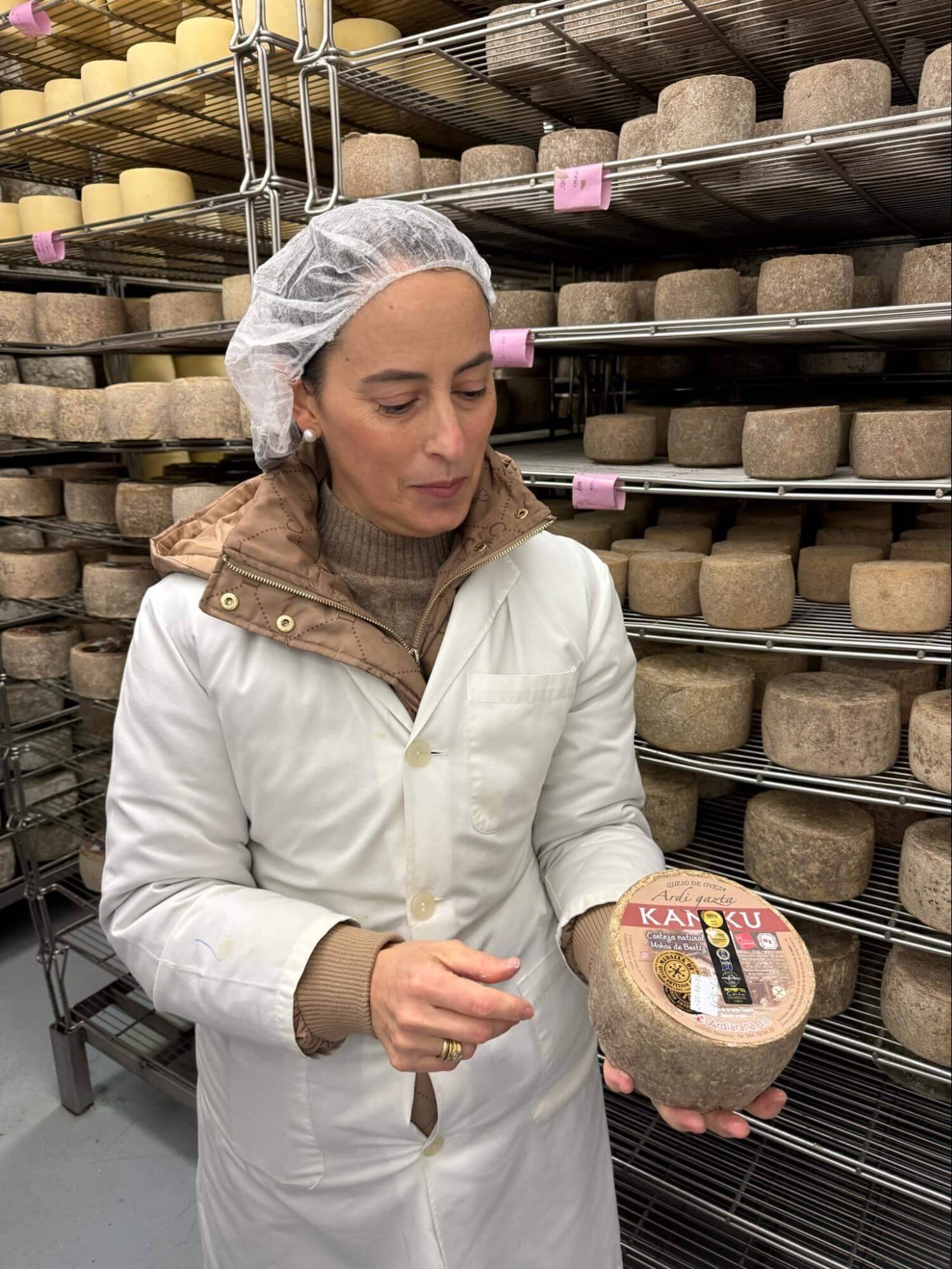 A person in a white lab coat and hair net holds a round cheese labeled 'KADOKU,' surrounded by shelves of various cheeses.