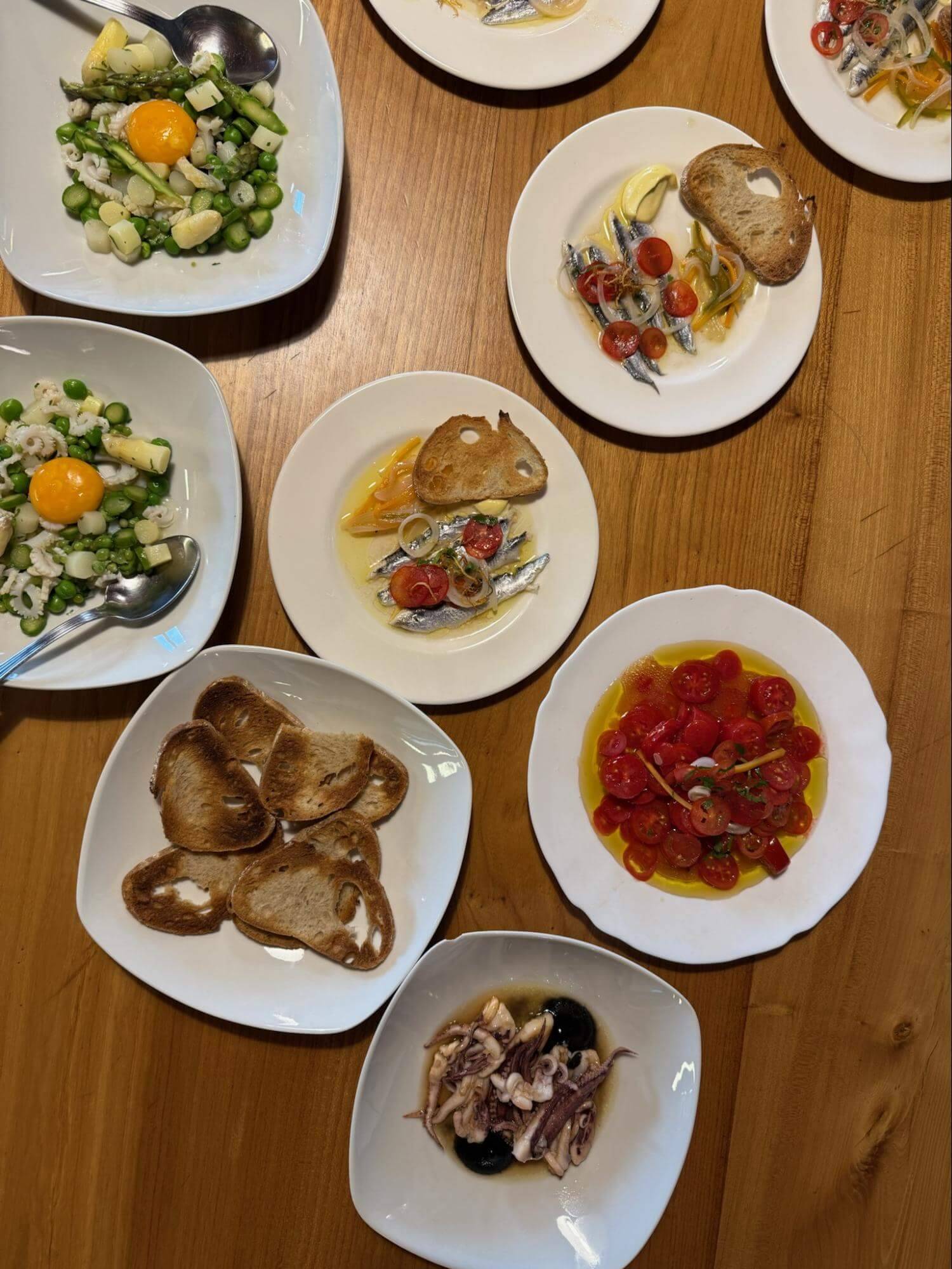 A variety of dishes on white plates, including salads, seafood, sliced bread, and cherry tomatoes, served on a wooden table.