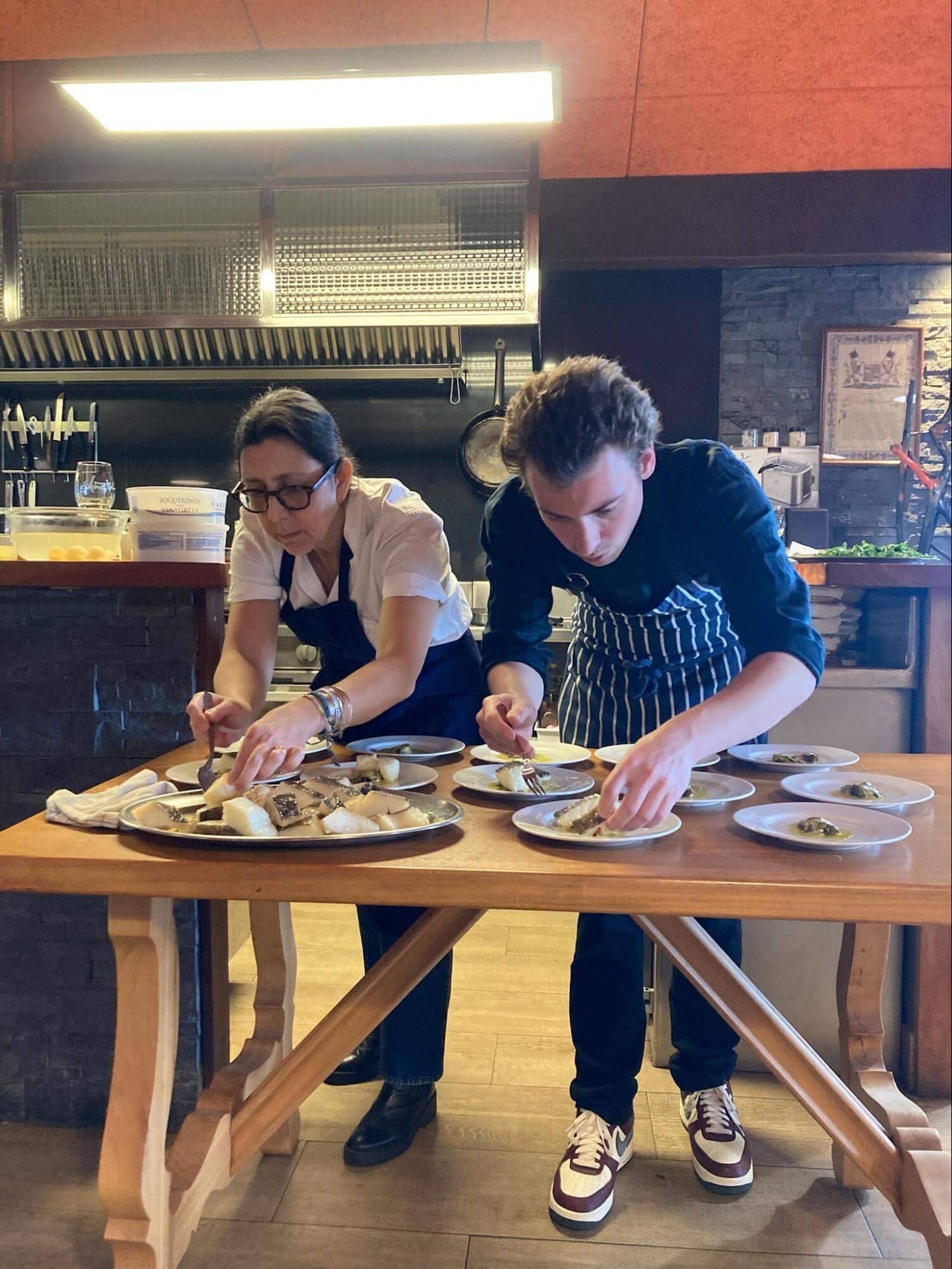 Two chefs are plating dishes at a wooden table in a professional kitchen, surrounded by plates and utensils.