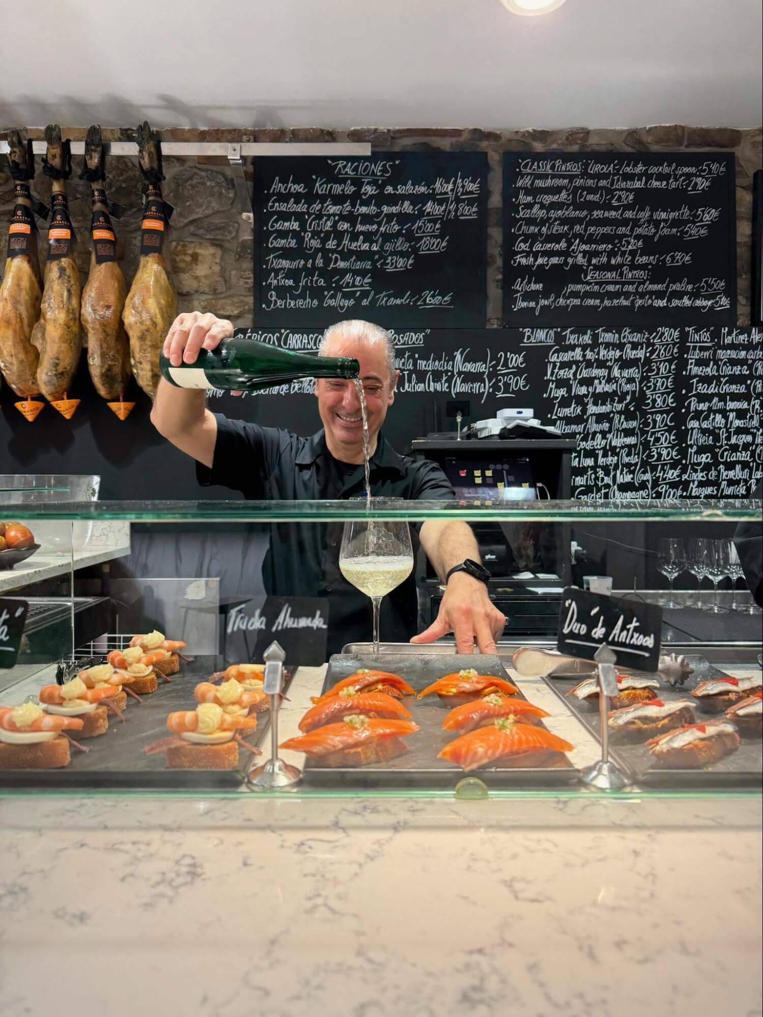 A restaurant counter displays a selection of gourmet tapas, with a bartender pouring a drink and jamón hanging in the background.