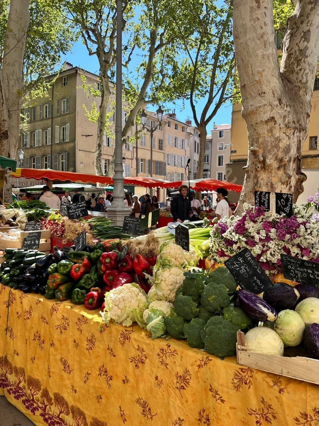 A vibrant marketplace filled with fresh vegetables, including bell peppers, cauliflower, and broccoli, under sunny skies and leafy trees.