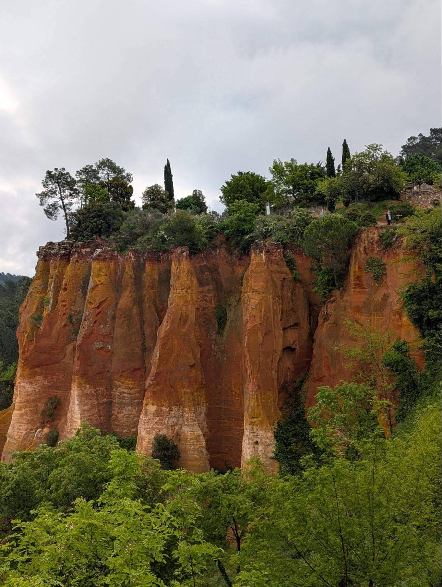 Vibrant orange cliffs rise above lush green trees under a cloudy sky, featuring scattered greenery and tall cypress trees.