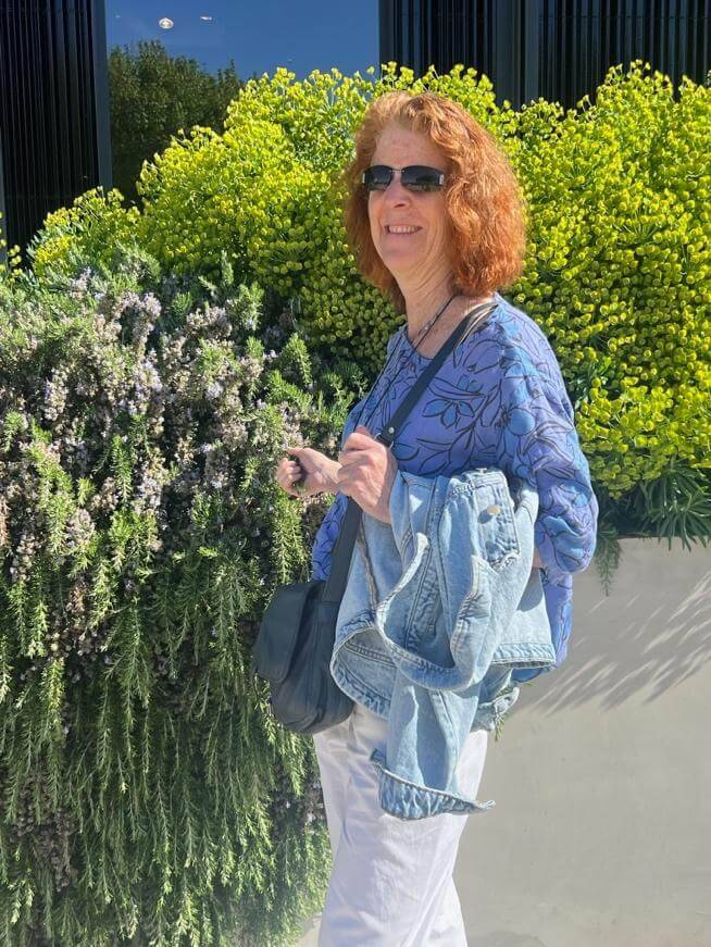 A woman stands against lush greenery, wearing a floral blue top and holding a denim jacket, with sunlight illuminating the scene.