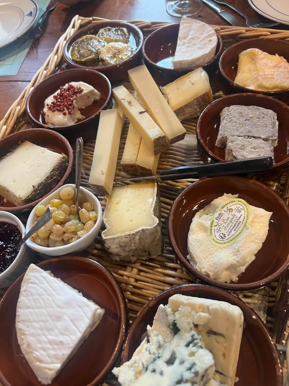 A wooden tray filled with assorted cheeses in bowls, accompanied by olives, jam, and garnishes, showcasing a rich variety of textures and flavors.