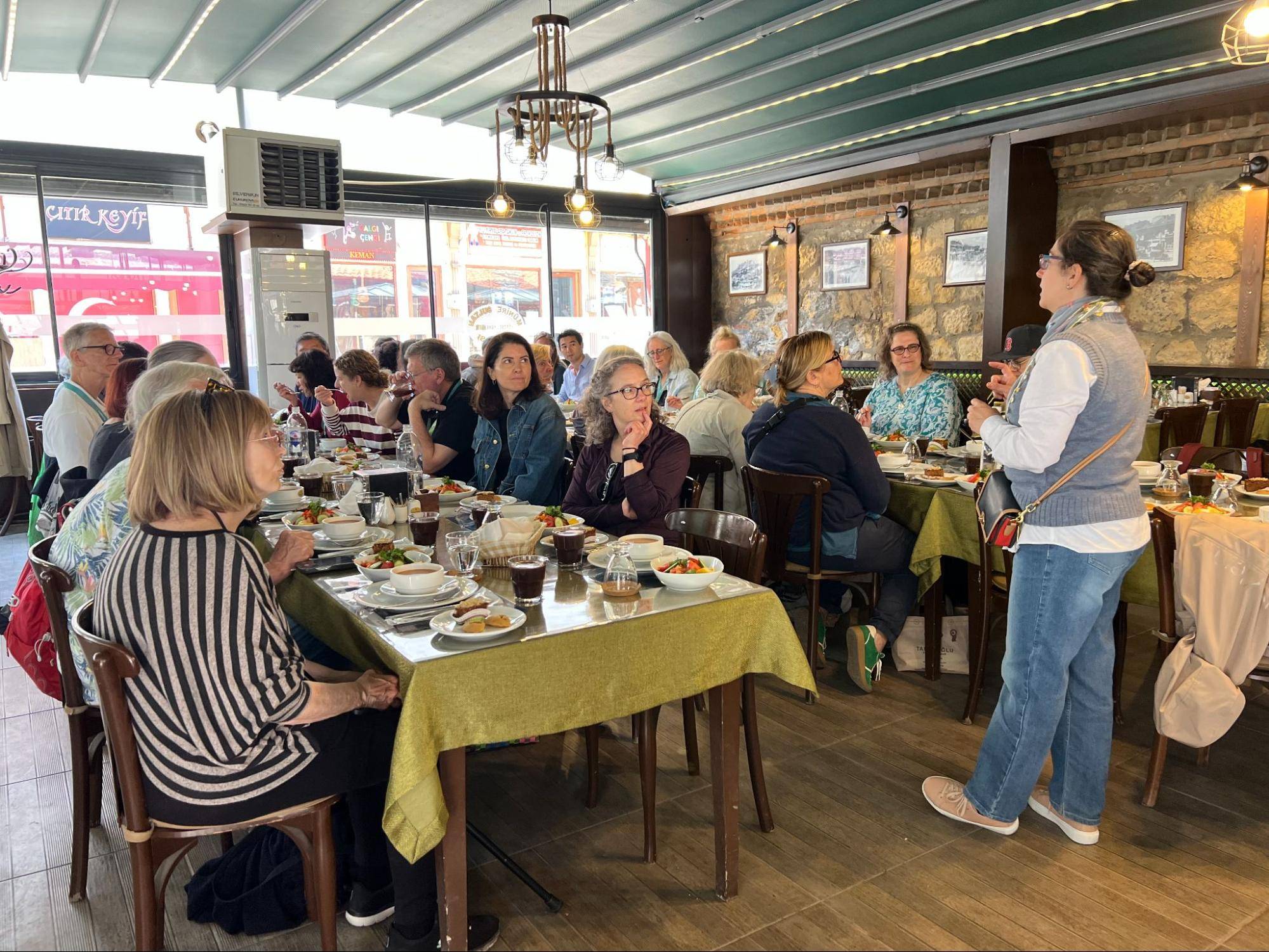 A group of people seated at a long table in a restaurant, enjoying a meal and engaging in conversation with plates of food in front of them.