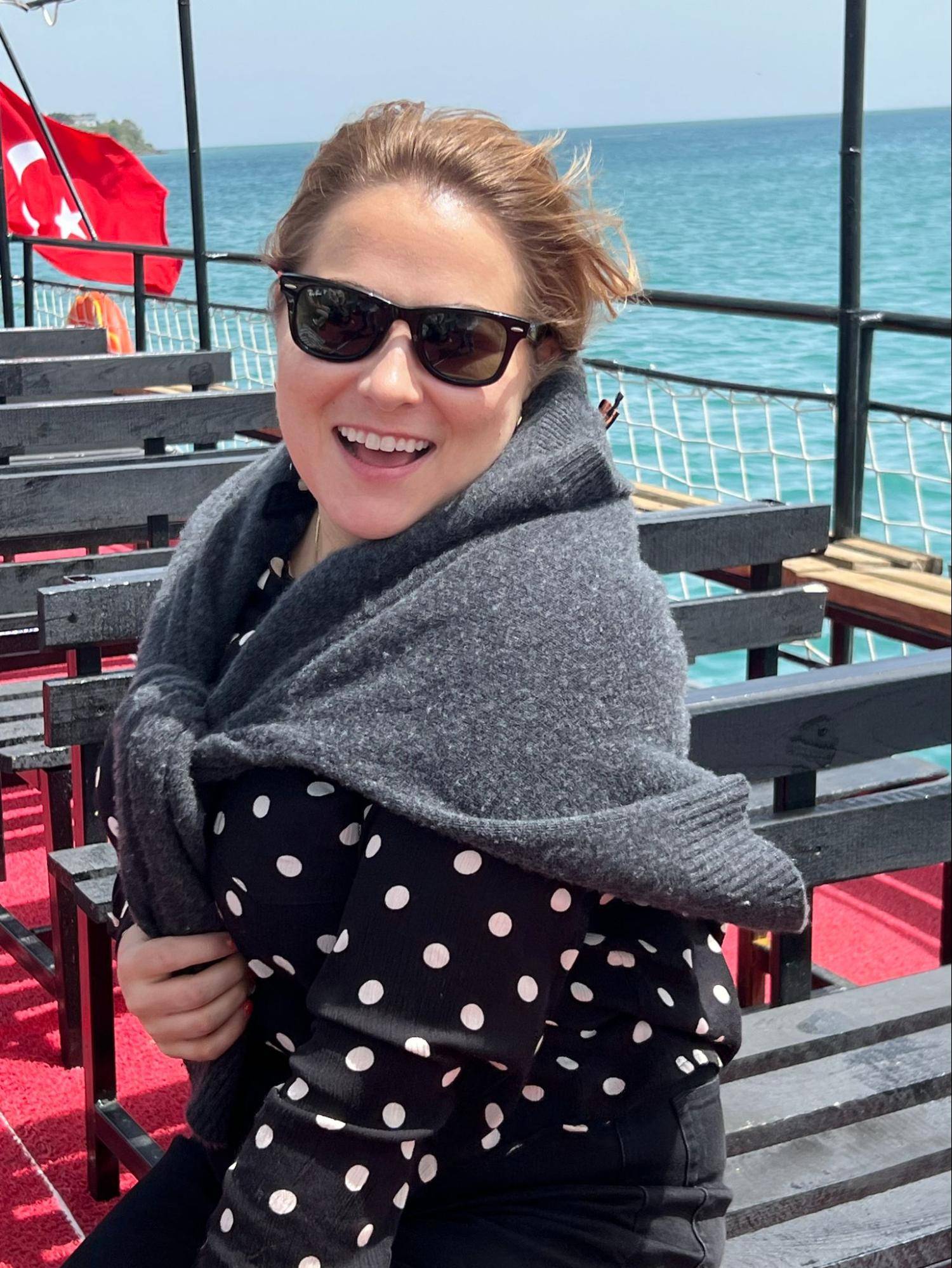 A person wearing a black polka dot shirt and a gray shawl sits on a boat deck, with a view of the sea and a Turkish flag in the background.