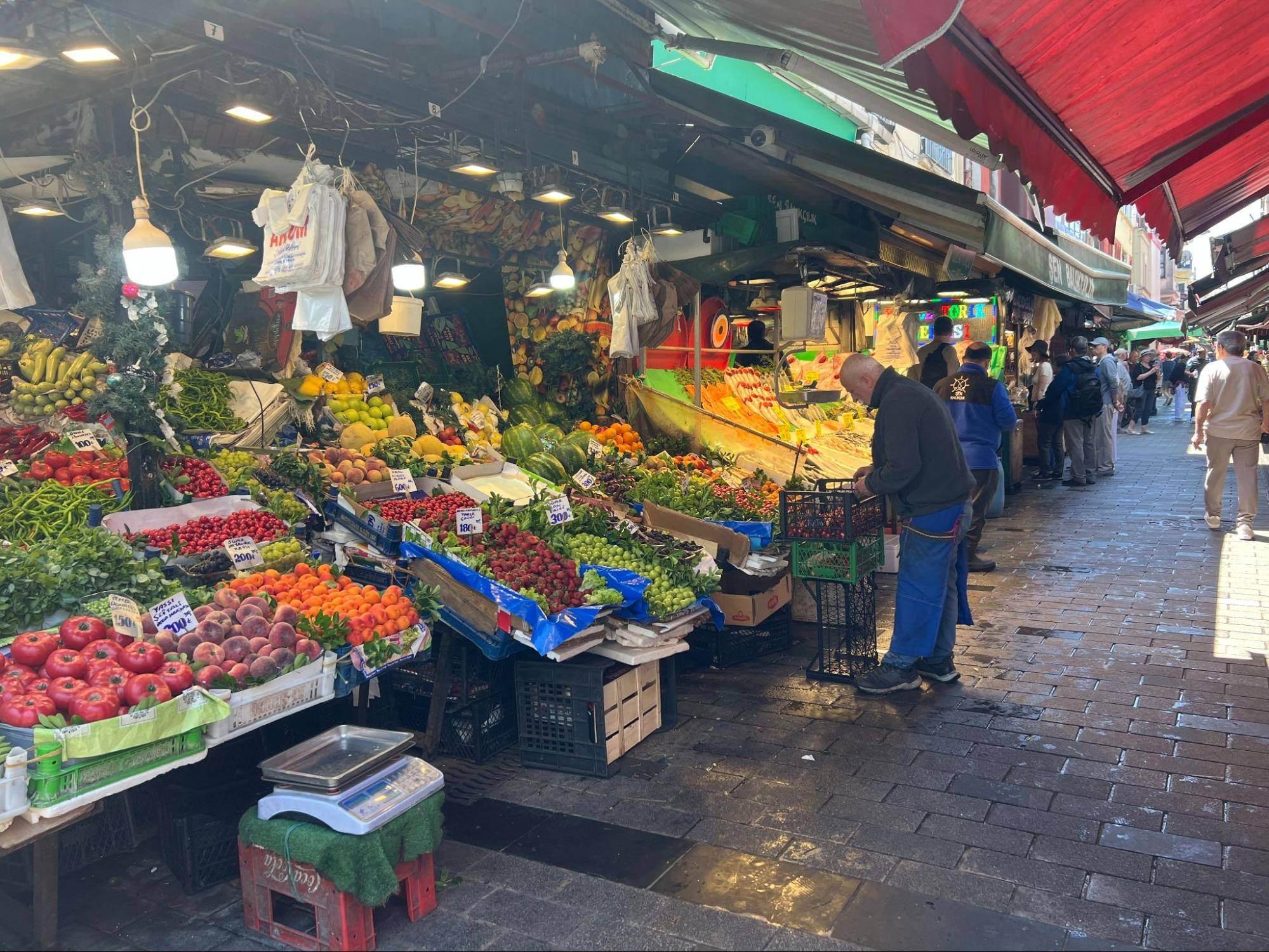 A vibrant market scene filled with colorful fruits and vegetables displayed on stalls, lush greenery, and shoppers browsing the aisles.