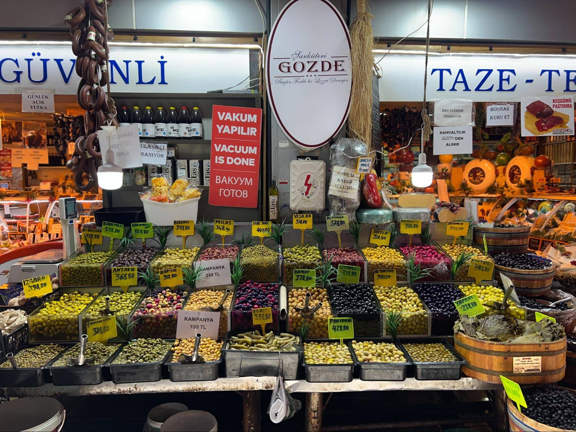 A vibrant market display featuring various types of olives in baskets, with price tags, and signs indicating vacuum sealing services.