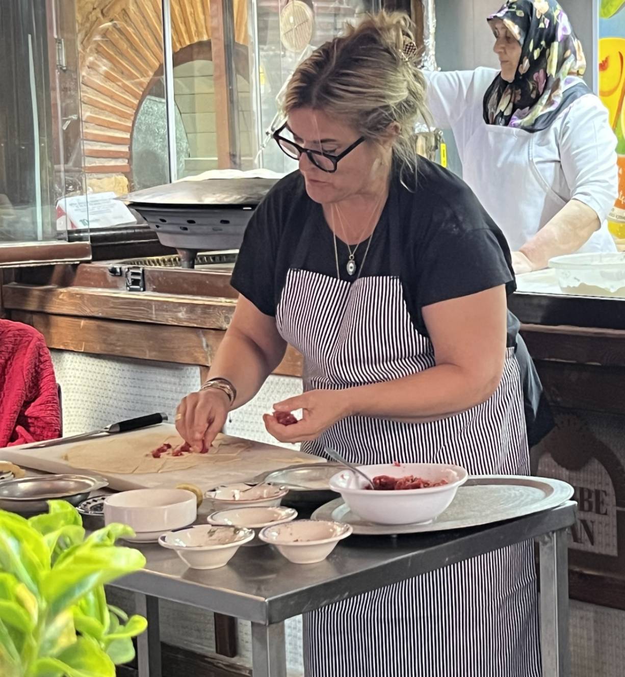 A woman in a striped apron prepares food at a table filled with bowls of ingredients, while another woman works in the background.