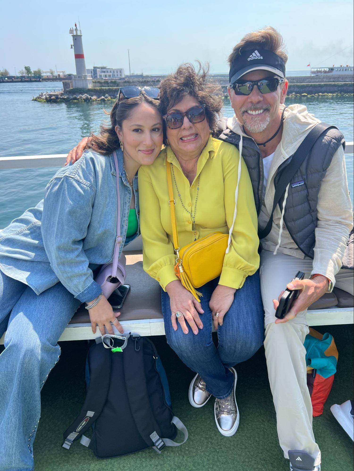 Three people sit together on a boat, with a lighthouse visible in the background and a sunny sky above. They are casually dressed.