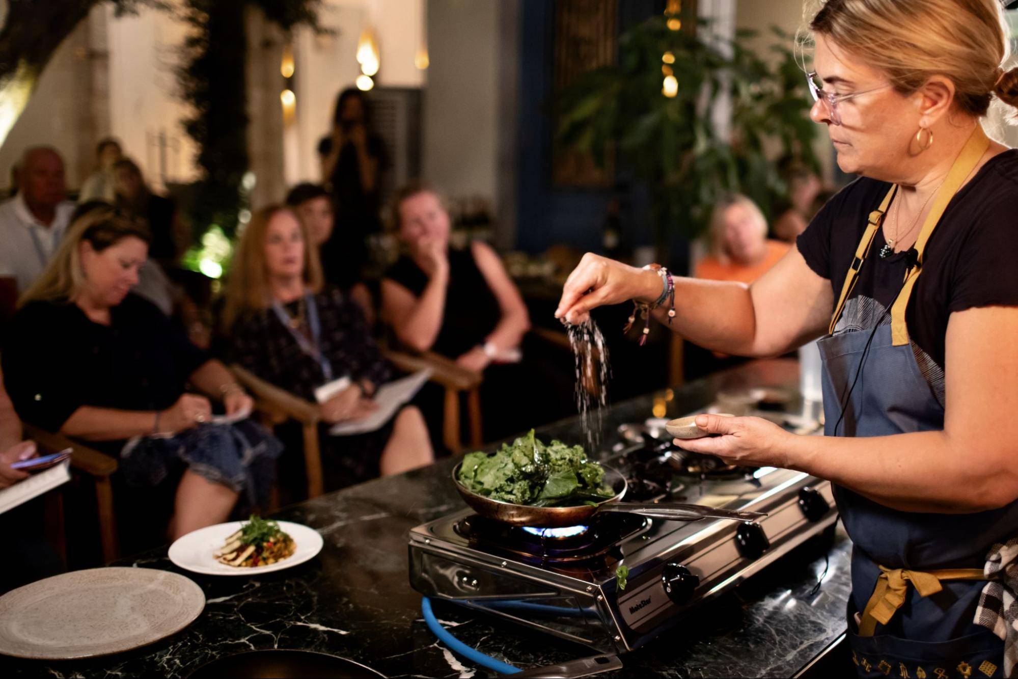 A chef adds seasoning to a pan of greens while a captivated audience watches attentively during a cooking demonstration.