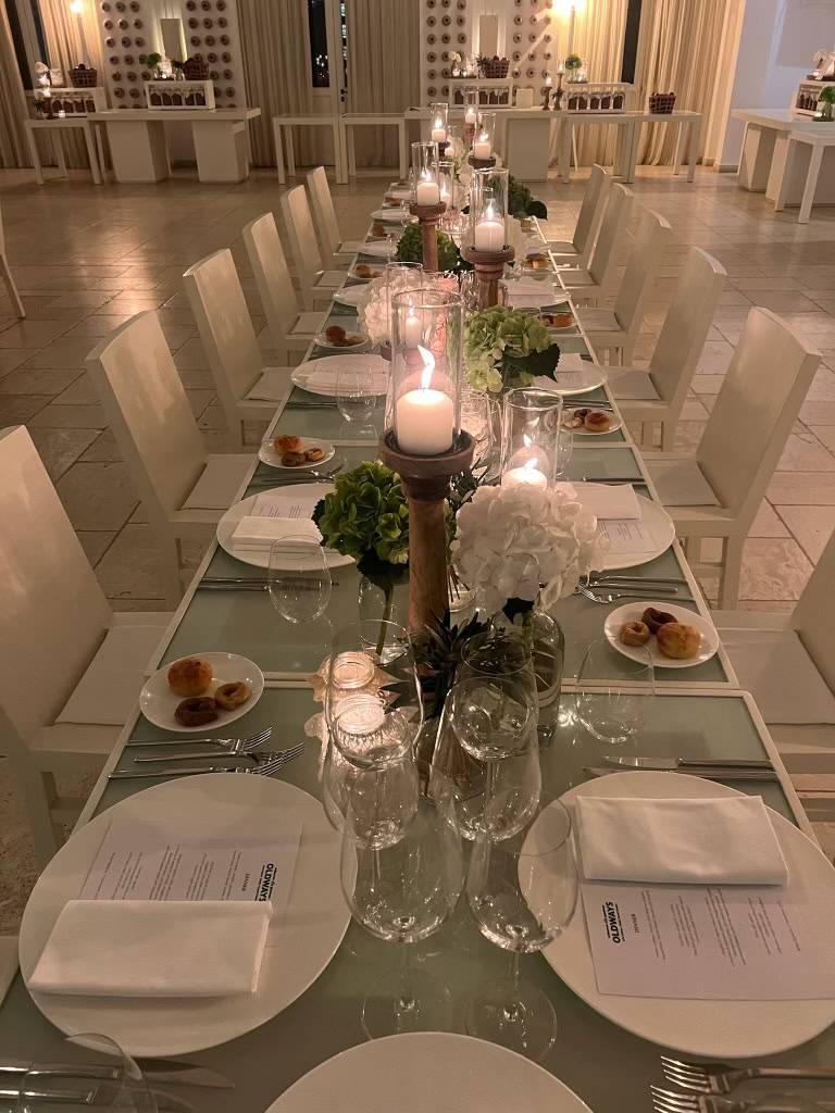 Elegant dining setup with white chairs, glass table, candles, hydrangeas, and place settings.