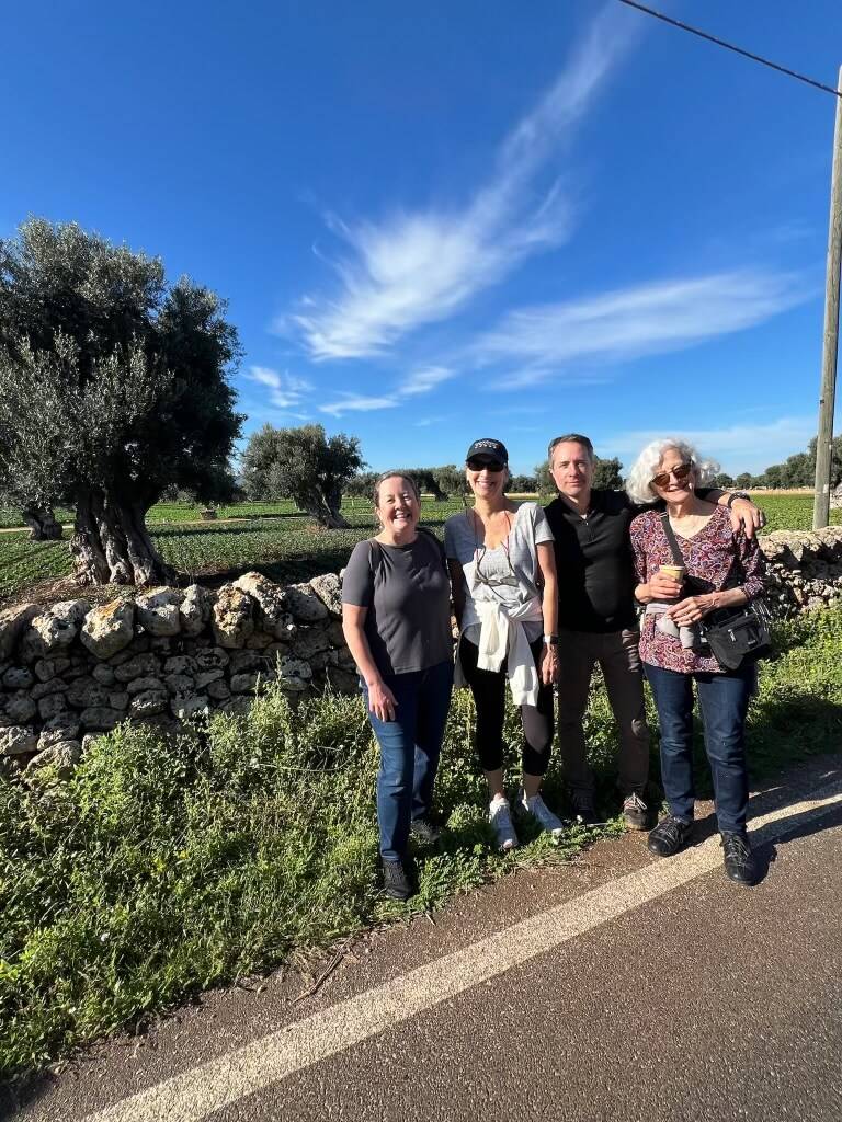 Four people pose by an old stone wall with olive trees and a clear blue sky.