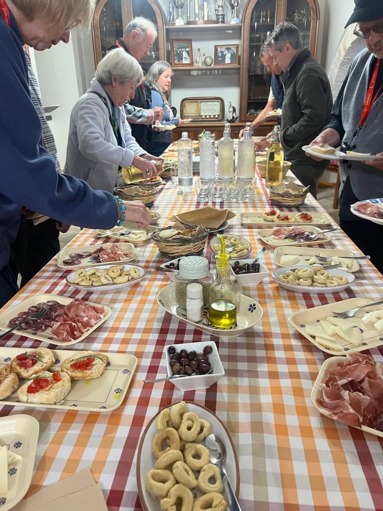 People serving themselves at an Italian buffet with meats, cheeses, olives, and breads.
