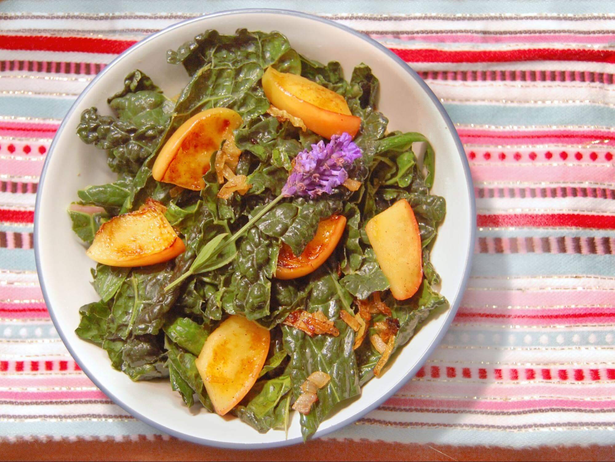 A vibrant salad with sautéed greens, caramelized apple slices, and a purple edible flower, served in a white bowl on a colorful striped tablecloth.