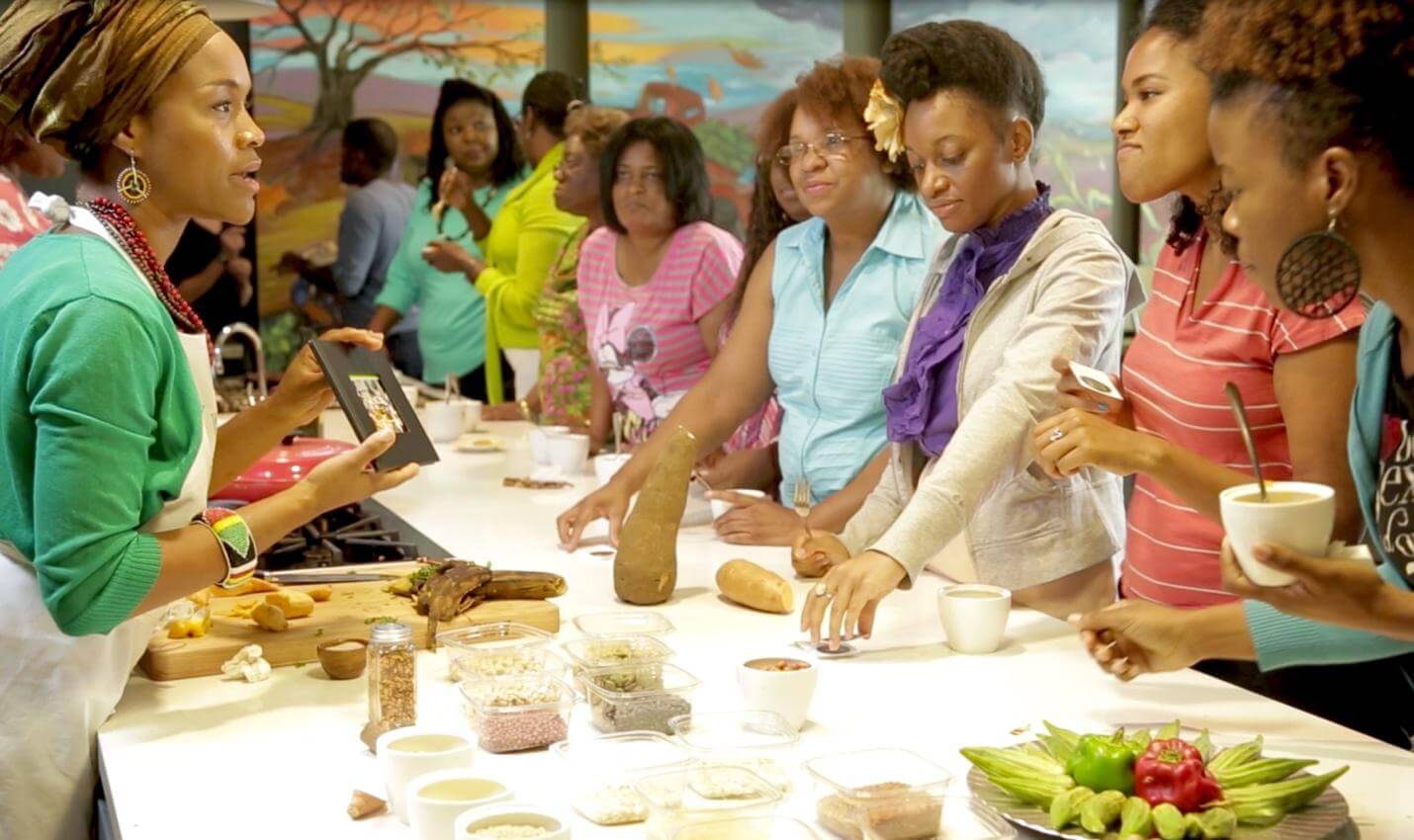 A diverse group of people gather around a kitchen island, engaging in food preparation and discussion, with ingredients displayed prominently.