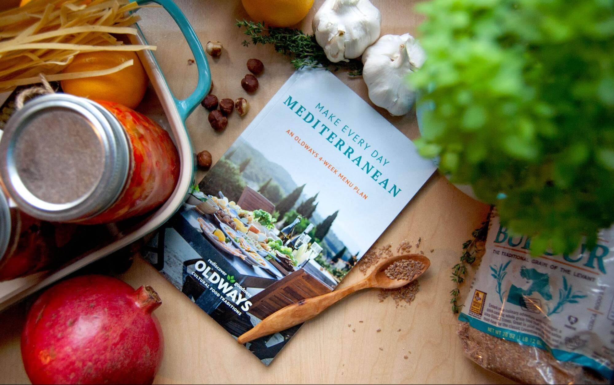 An assortment of Mediterranean cooking ingredients surrounds a cookbook titled 