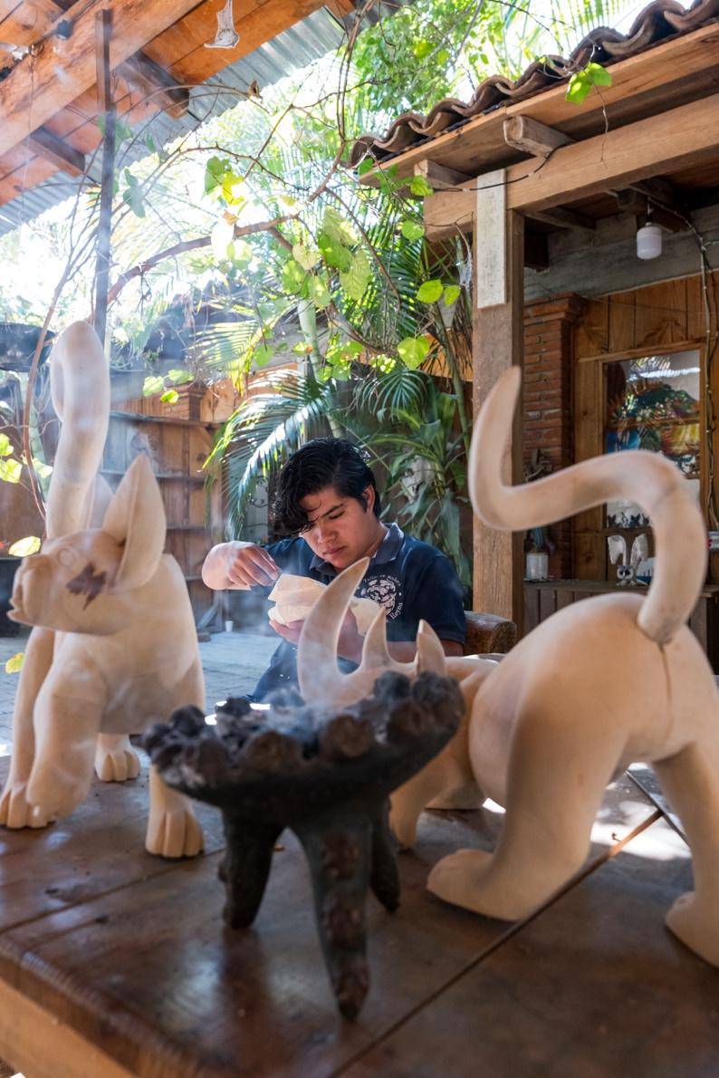 A woman sculpting a cat from clay, focused on her work in a well-lit studio filled with art supplies.