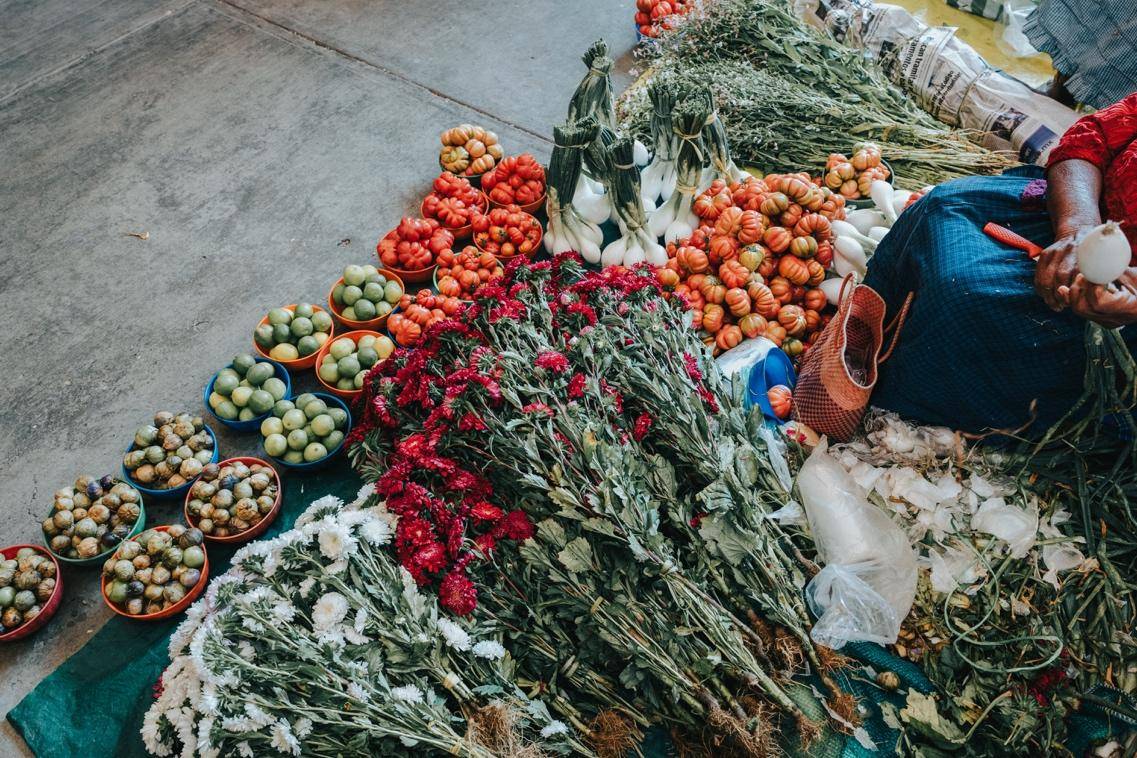 A woman sits on the ground surrounded by a colorful bunch of flowers, smiling gently as she admires them.