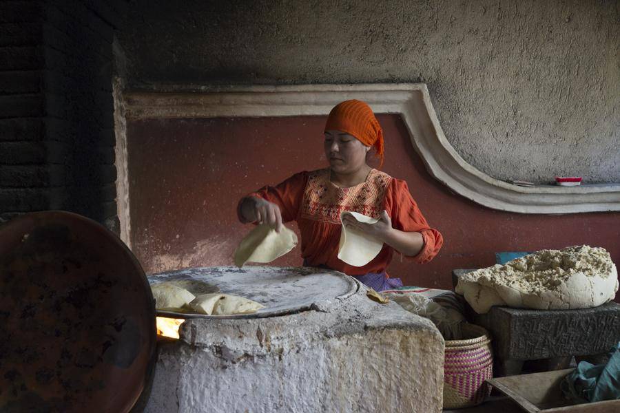 A woman wearing an orange turban kneads dough while making bread in a cozy kitchen setting.