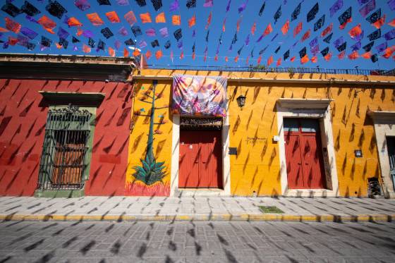 Vibrant streetscape featuring colorful buildings, red doors, and papel picado decorations fluttering against a clear blue sky.