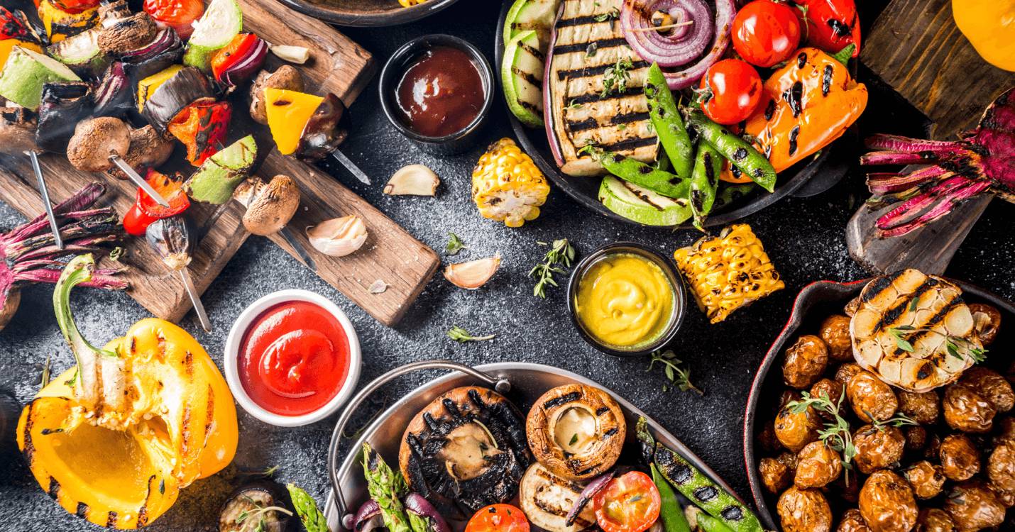 A vibrant spread of grilled vegetables, skewers, and sauces arranged on a dark surface, showcasing colorful peppers, corn, and mushrooms.