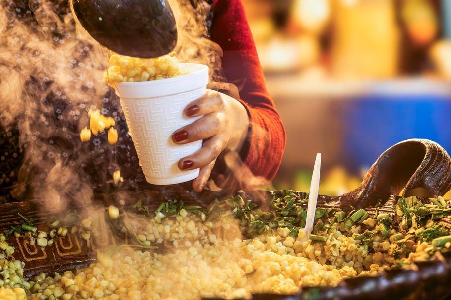 A hand with red nails scoops steaming corn into a white cup