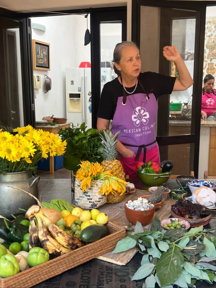 A woman in a purple apron gestures animatedly in a kitchen