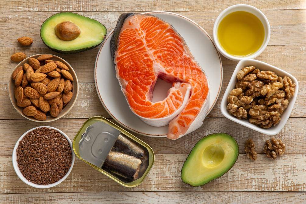 Assorted healthy fats on a wooden table: salmon steak, avocados, almonds, walnuts, flaxseeds, sardines, and olive oil.