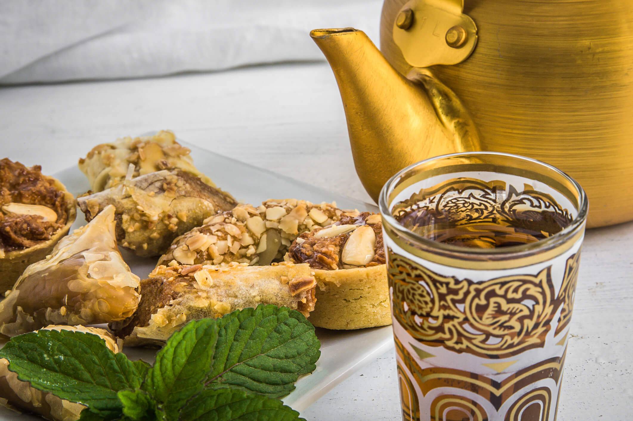 A plate of assorted nut-topped pastries, a gold teapot, a decorative glass of tea, and mint leaves on a white table.