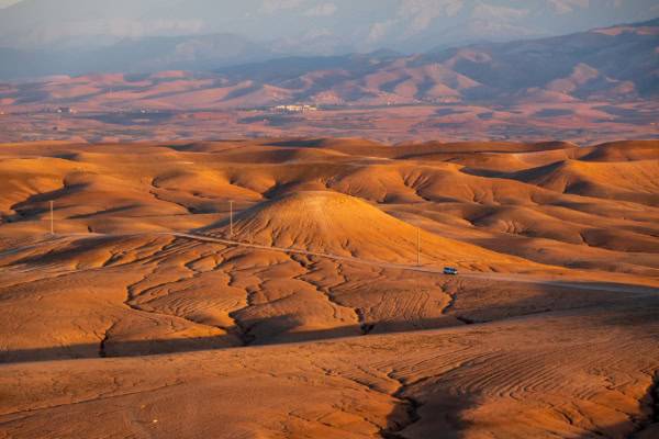 Expansive desert landscape with rolling hills, dirt road, and distant mountains under a clear sky during sunset.